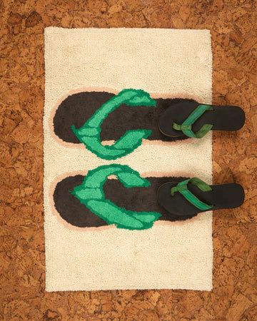 A pairs of black and green flip-flops on a textured bath mat with flip flop design on a cork-like background.