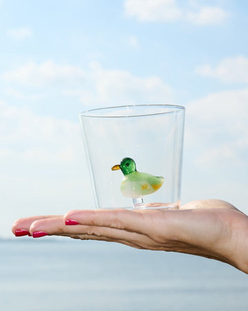 A small cup with a duck figure made of glass inside sits in the palm of a person's hand. The person has pink pained nails. 