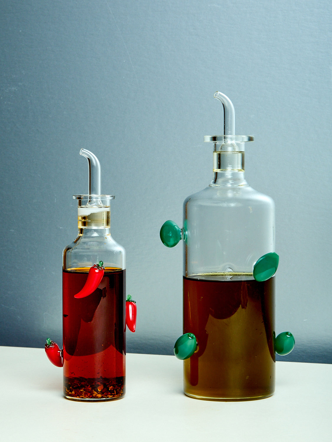 Two glass bottles, one filled with red chili oil and the other with olive oil, each adorned with decorative olives and peppers.