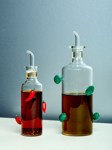 Two glass bottles, one filled with red chili oil and the other with olive oil, each adorned with decorative olives and peppers.