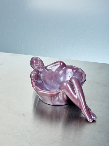 Plum purple glass sculpture of a reclining figure on a reflective surface with a light blue background