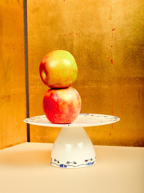 Two apples stacked on a small white cake stand against a gold background