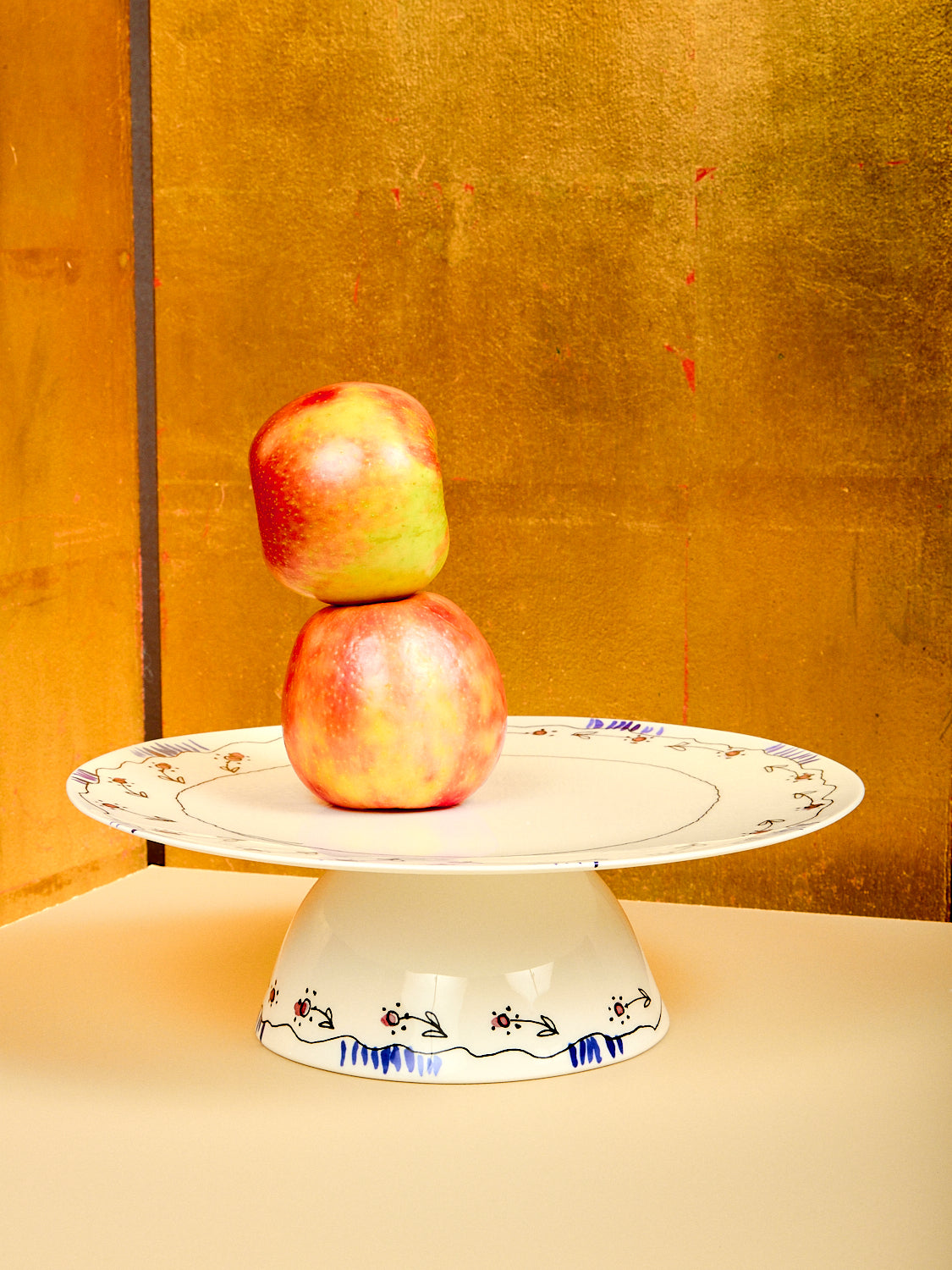 Two apples stacked on a decorative white cake stand against a gold background