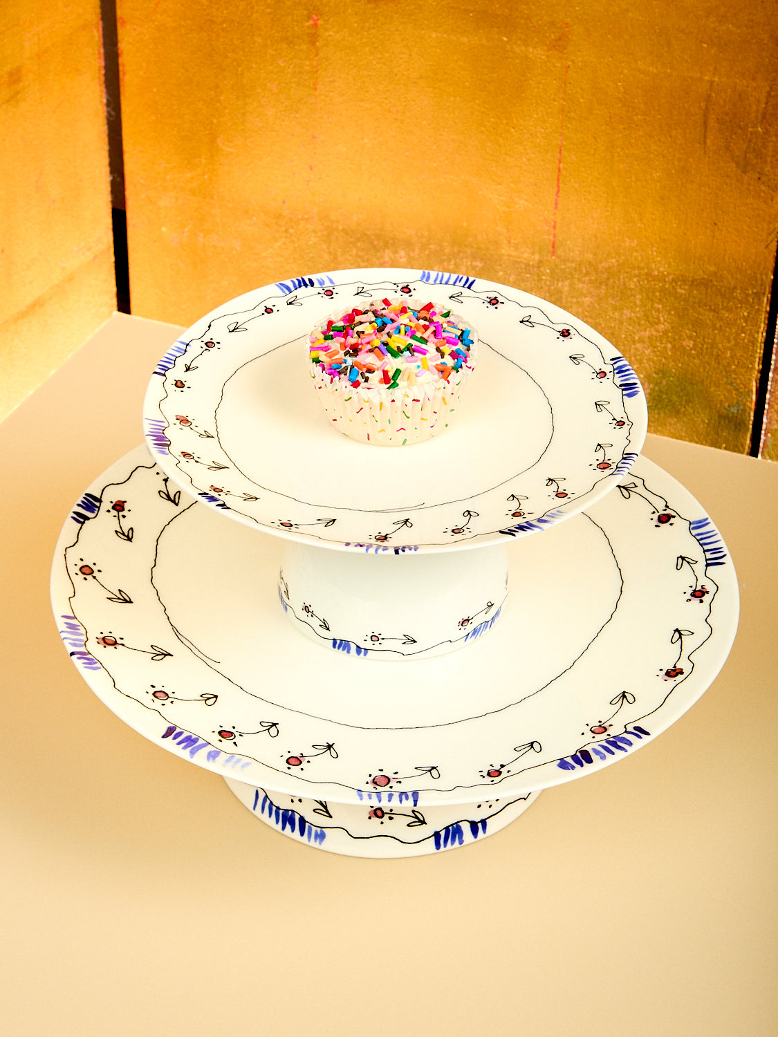 Two stacked decorative cake stands with a small cake on a gold background
