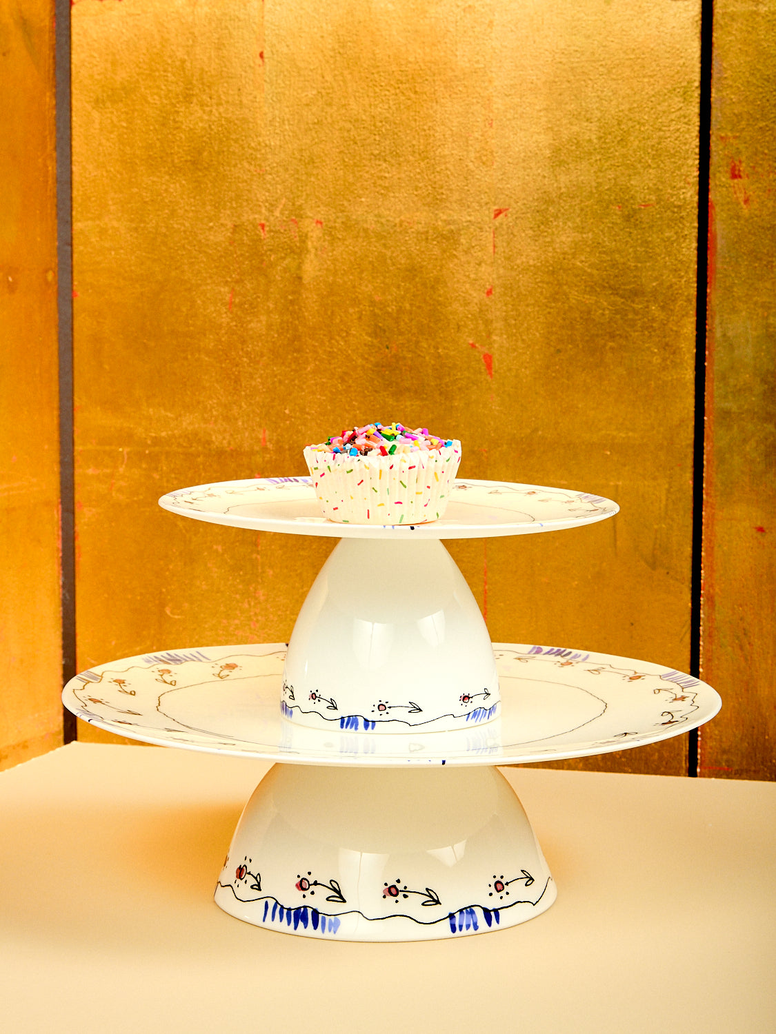 Two stacked ceramic cake stands with a small cake on a gold background