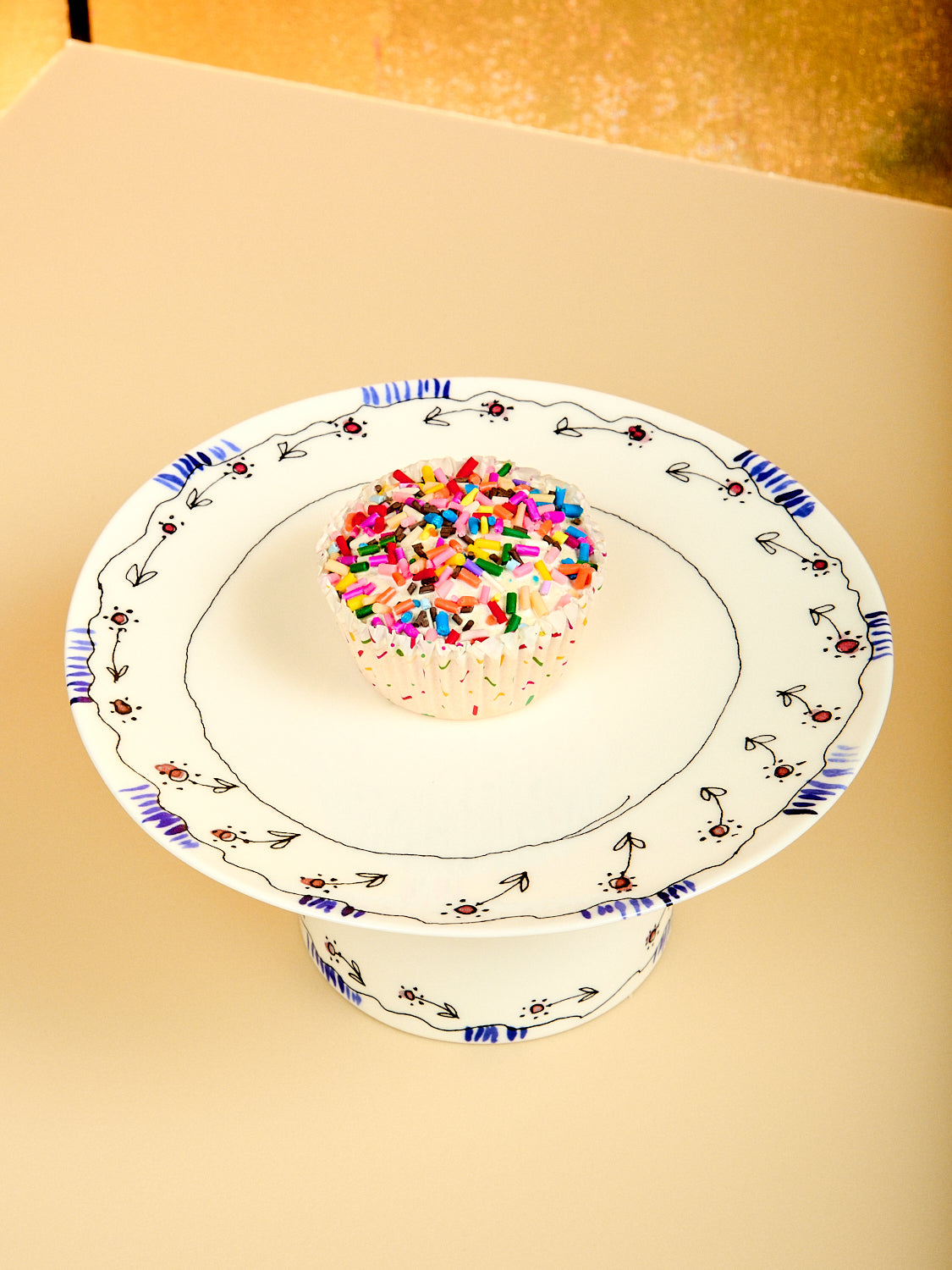 Small cake with colorful sprinkles on a decorative white plate with blue floral patterns against a beige background.