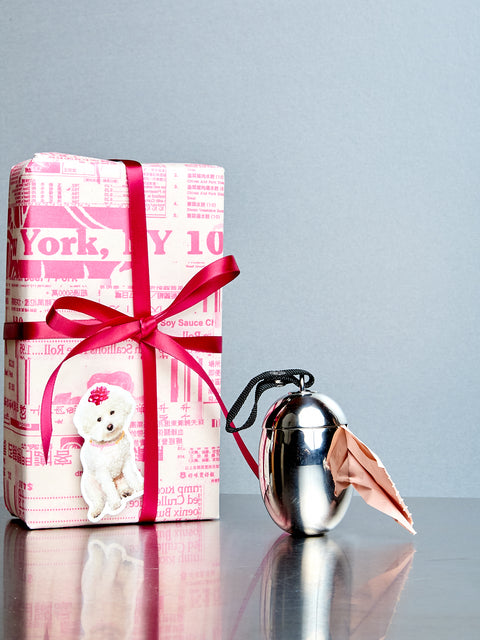 Pink wrapped gift with a red ribbon and a silver dog waste bag holder on a reflective surface.
