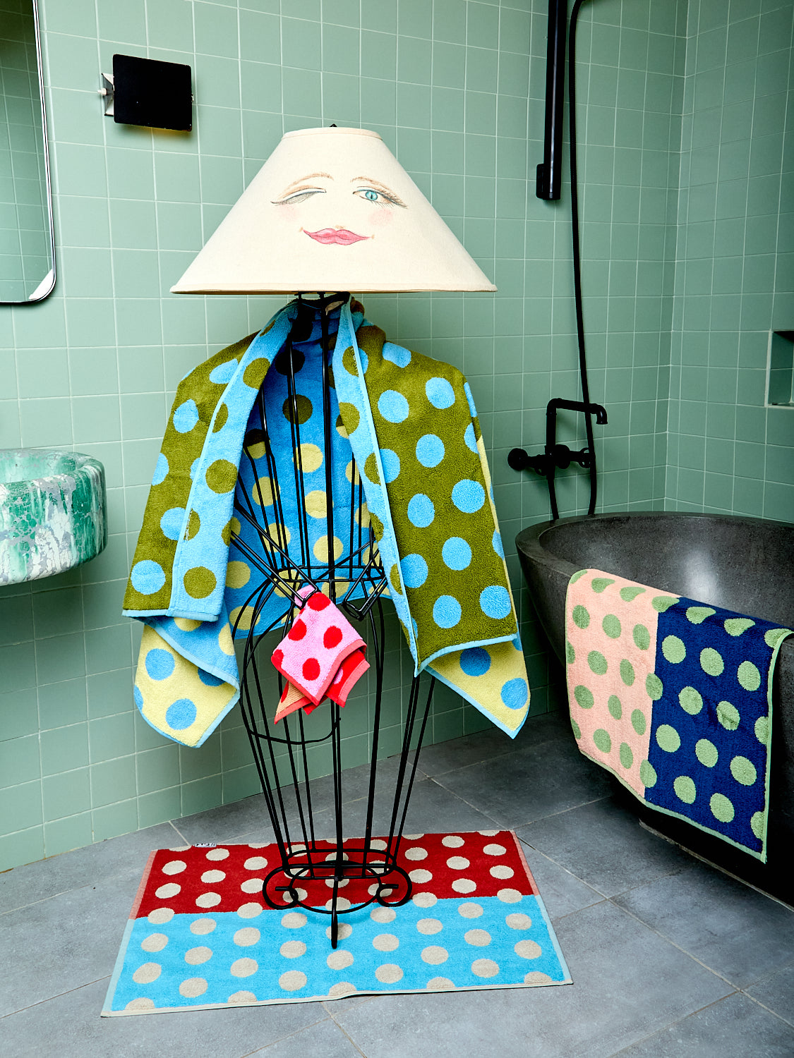 Two colorful polka dot towels, a hand towel and bathmat, green tiled walls, and a bathtub.
