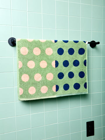 Green bath towel with pink and blue polka dots hanging on a rack against a light green tiled wall.