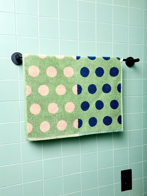 Green bath towel with pink and blue polka dots hanging on a rack against a light green tiled wall.