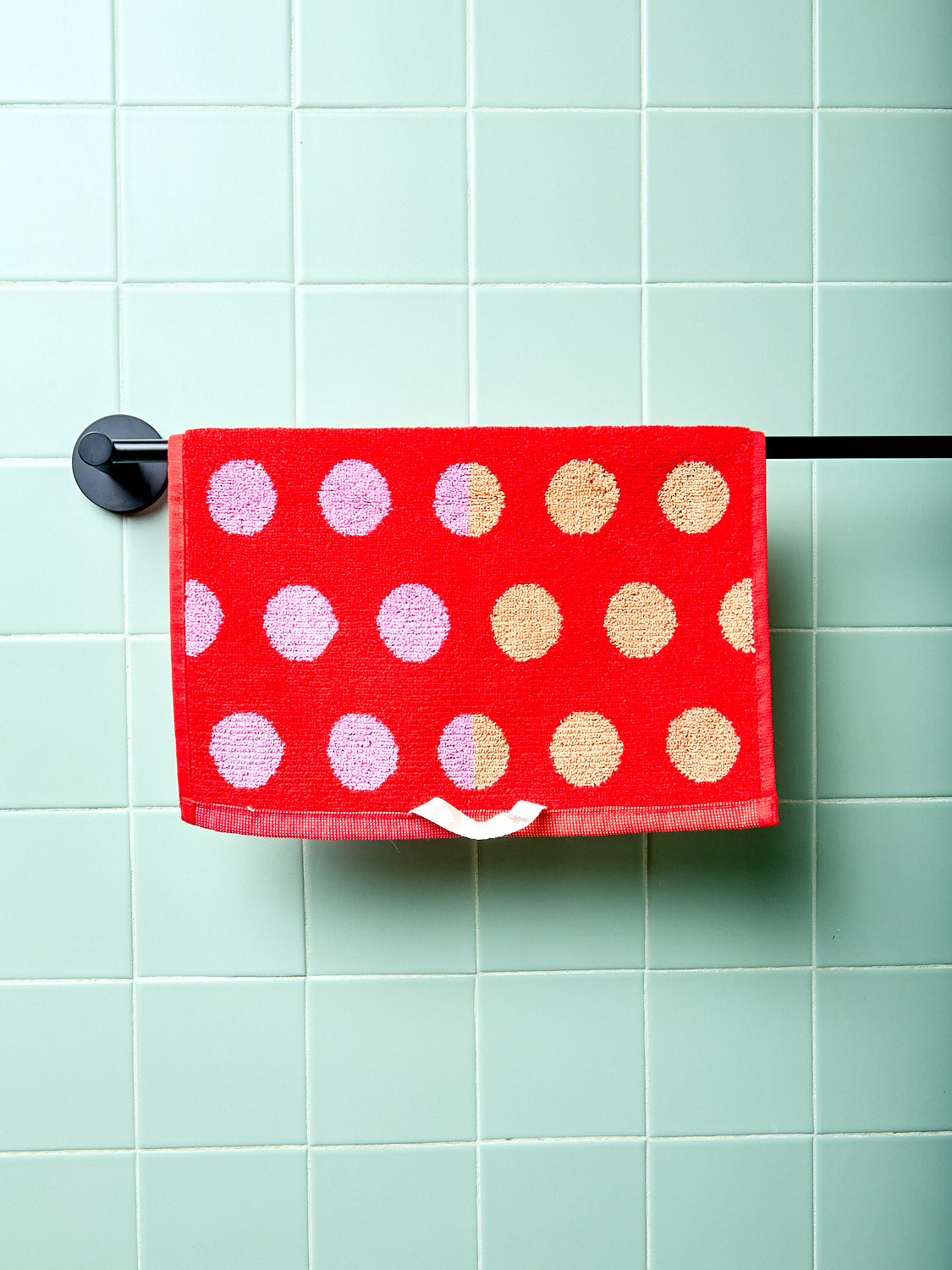 Red hand towel with pink and gold polka dots hanging on a rack against a light green tiled wall.