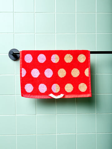 Red hand towel with pink and gold polka dots hanging on a rack against a light green tiled wall.
