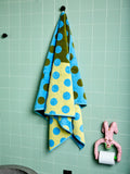 One green and yellow bath towel with blue polka dots hanging on a tiled wall next to a pink toilet paper holder.